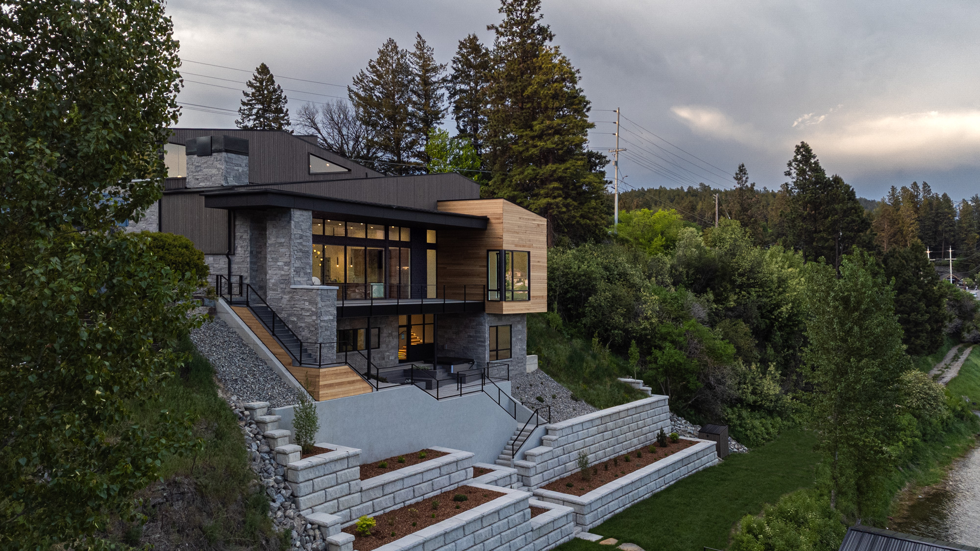 Modern multi-level house with large windows, stone and wood exterior, built on a hillside surrounded by trees.