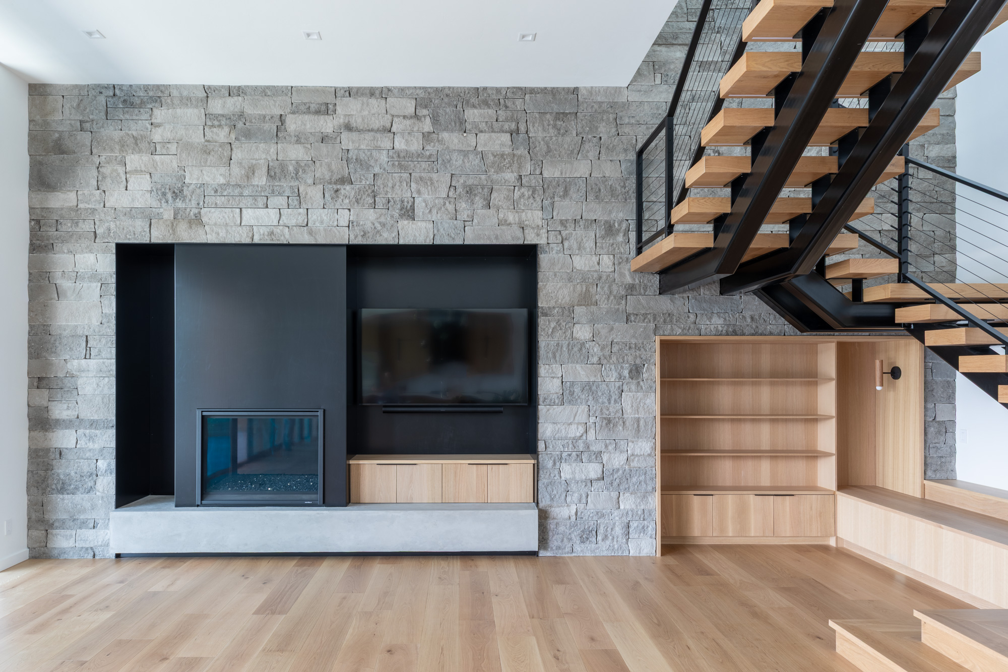 Modern living room with stone accent wall, TV, fireplace, wooden shelves, and open wooden staircase with black railing.
