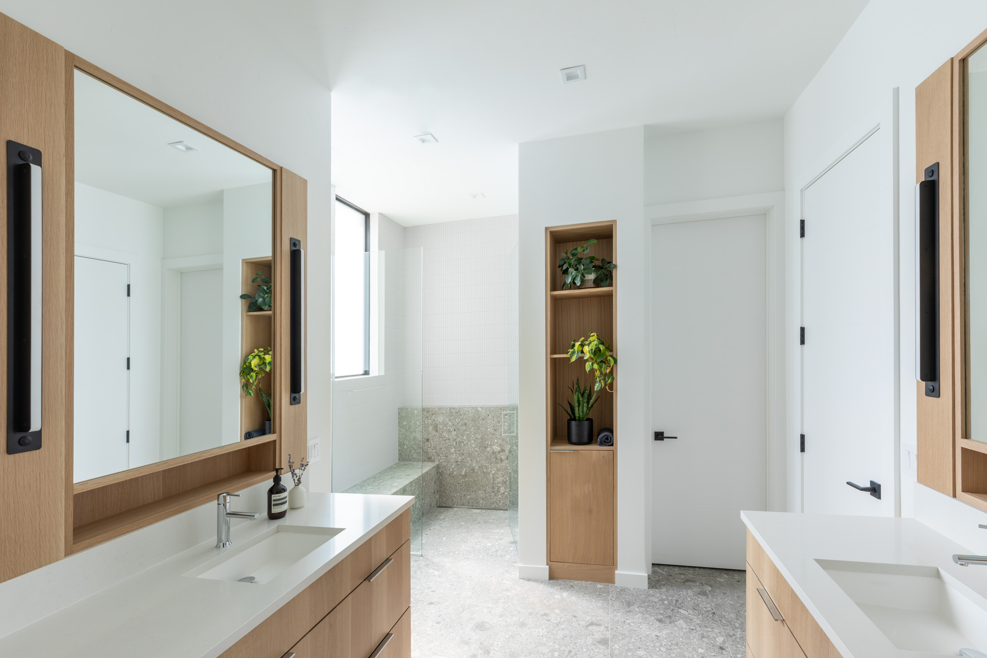 Modern bathroom with light wood cabinets, white countertops, large mirrors, plants, and a walk-in shower with a bench.