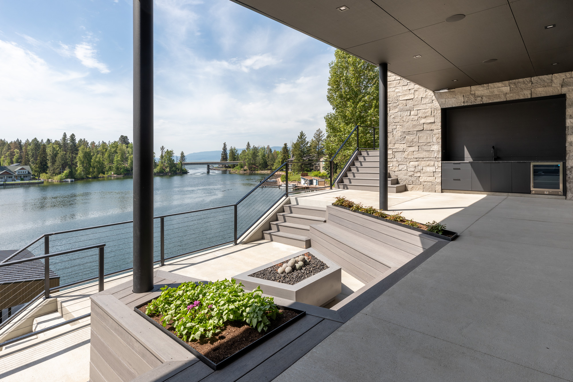 Modern lakeside patio with built-in seating, planters, and a fire pit, overlooking water and trees under a clear sky.