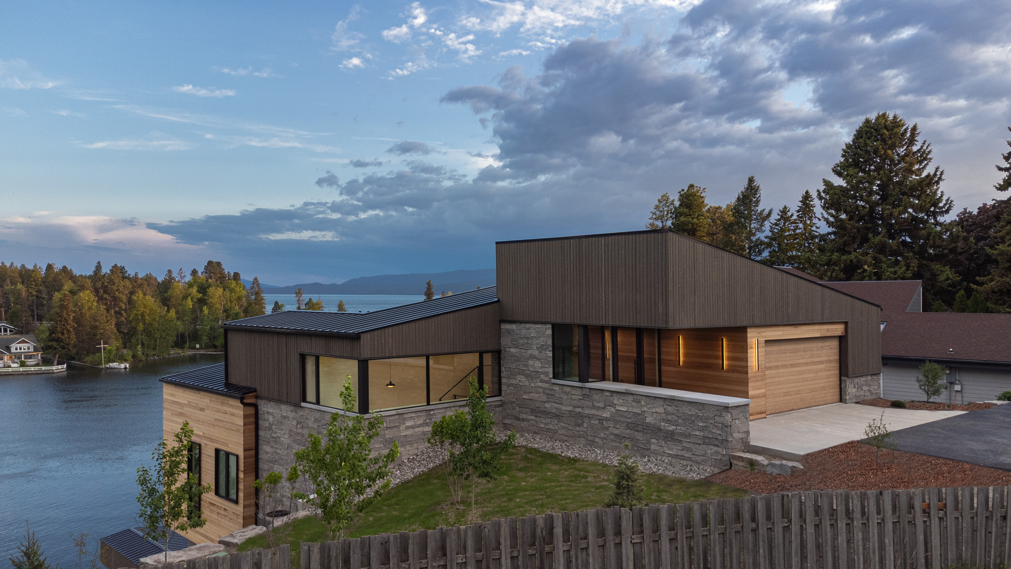 Modern hillside house with wood and stone siding, beside a calm lake and a wooden fence
