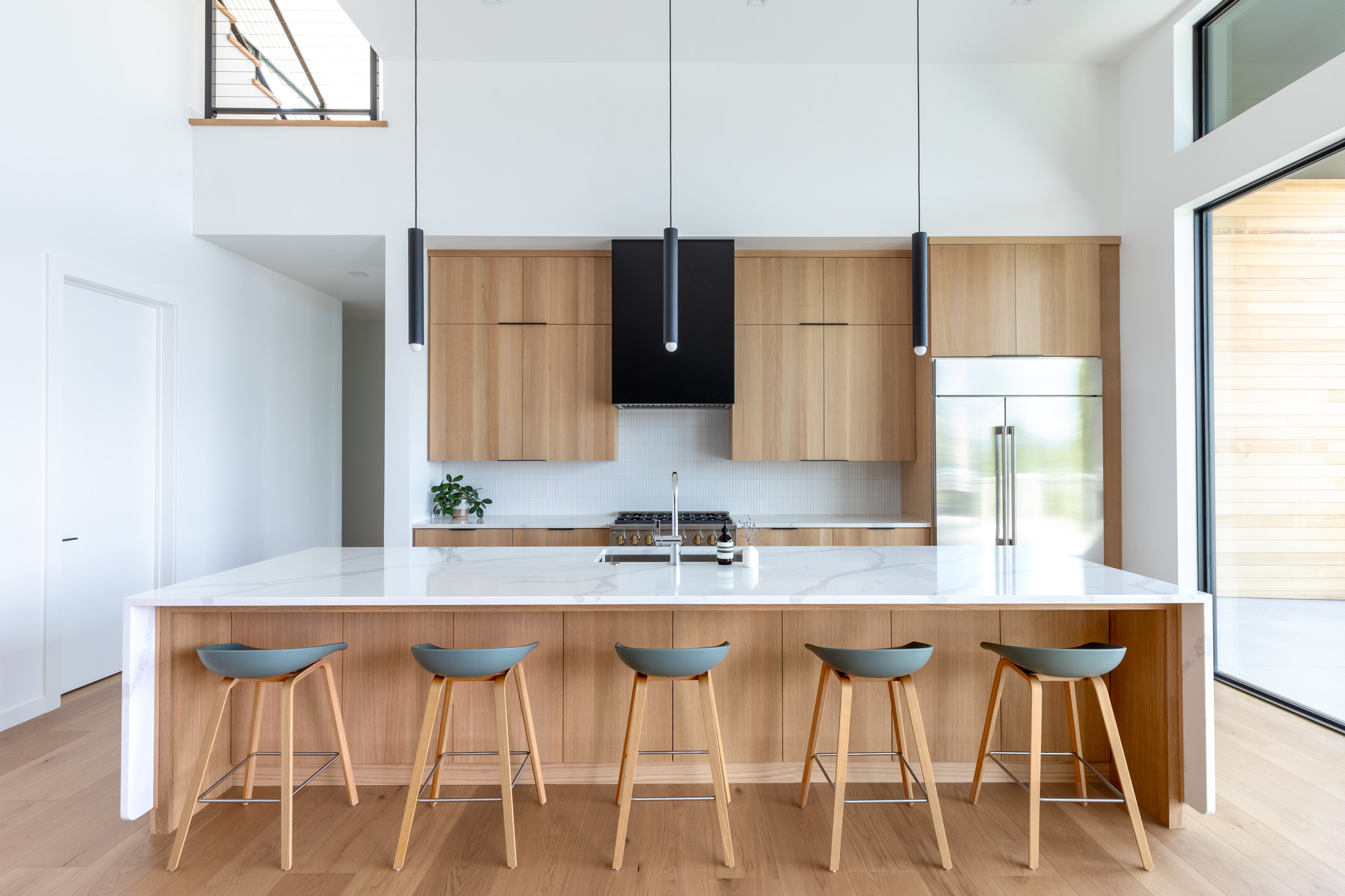 Modern kitchen with a large white island, wooden cabinets, and a stainless fridge against the wall; pendant lights hang over the island.