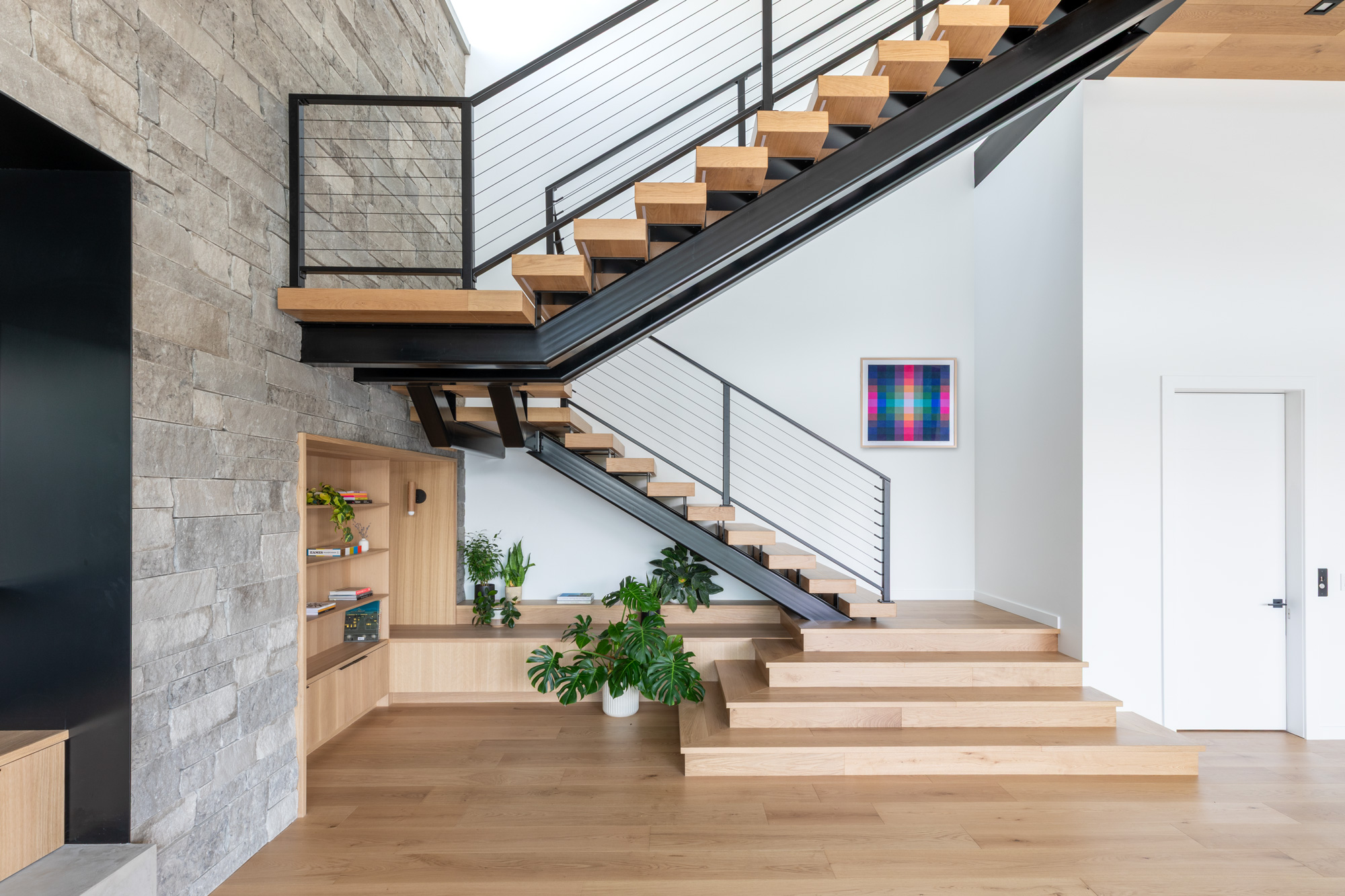 Interior living area with a floating wooden staircase and black metal railing beside a stone wall; green plants and a colorful framed artwork on the white wall.